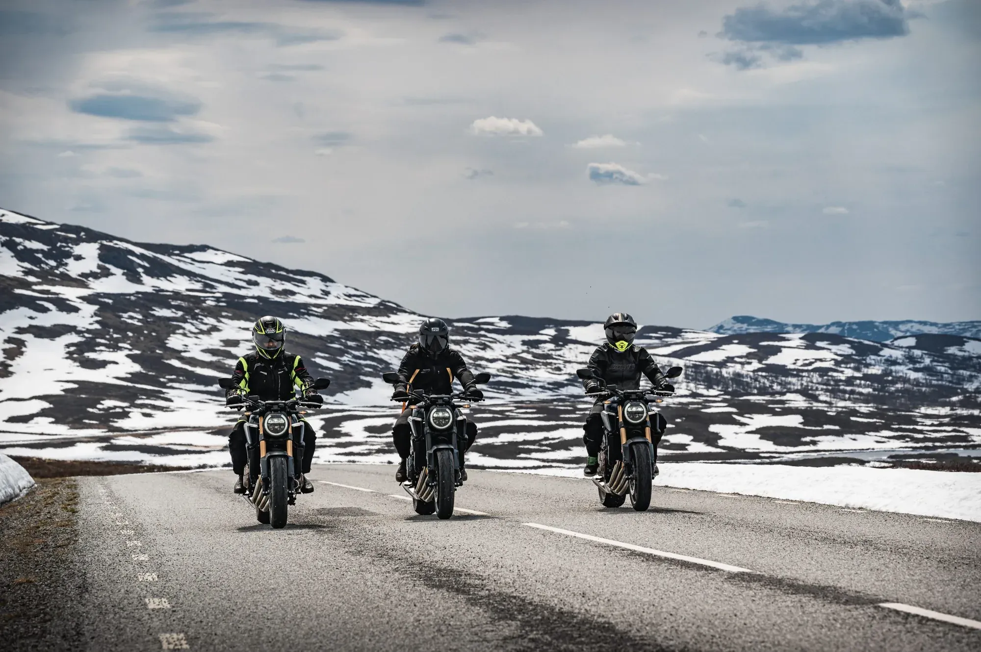 Tre motorcyklister på en fjällväg med snöklädda berg i bakgrunden
